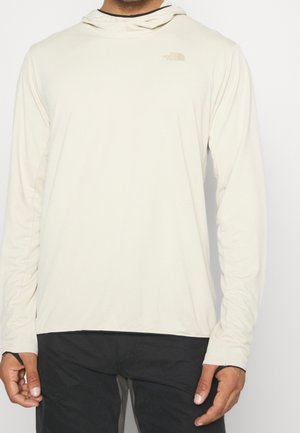 Mies, jolla on pitkähihaiset beige-huppari, jossa on peukaloaukot, yhdistettynä mustiin housuihin, seisoo yksinkertaisen valkoisen taustan edessä.