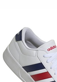 Bijele kožne tenisice s teksturiranom završnom obradom, koje imaju mornarsko plave i crvene pruge. Uključuju bijele vezice i podstavljeni ovratnik s "adidas" logotipom.