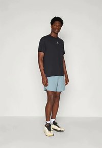 Must värvi meeskonna kaelusega T-särk väikese logoga, helesinised lühikesed püksid ja must-valged spordijalatsid. Lühikesed tumedad juuksed, seistes.