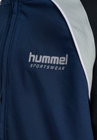 Navyblaue Sportjacke mit hellgrauen und schwarzen Einsätzen, mit "hummel SPORTSWEAR" Logo auf der Brust.