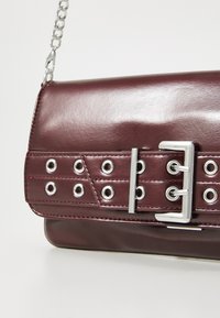 Sac à main en simili cuir bordeaux avec une finition brillante, présentant des œillets en argent, une accentuation en boucle et une bandoulière en chaîne. Forme rectangulaire.
