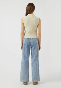 Top sin mangas de punto beige con dobladillo en forma de concha, combinado con jeans de talle alto y pierna ancha de color azul claro y calzado beige.