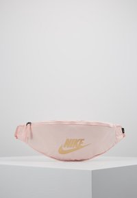 Růžová látková ledvinka s zakřiveným designem, na které dominuje výrazné zlaté logo Nike. Má zipové zapínání a nastavitelný popruh.