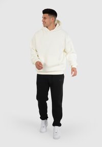 Cremefarbener Kapuzenpullover mit einer Fronttasche, kombiniert mit schwarzen Jogginghosen und weißen Sneakern. Lässiger Schnitt mit weicher Textur.