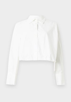 Kuttet hvit langermet skjorte med knapper, krage og justerbar snor i kanten på ensfarget bakgrunn.