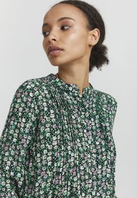 Blusa floreale in verde scuro con fiori neri, bianchi e rosa. Presenta una parte anteriore plissettata e un colletto alto senza colletto. Tessuto leggero.