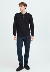 Nuori mies, jolla on yllä musta pitkähihainen poolopaita, tummansiniset farkut ja mustat kengät, seisoo vaaleaa taustaa vasten.