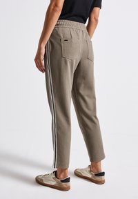 Persoon in taupe tapered broek met witte zijstrepen en achterzakken, staand, gecombineerd met beige en witte sneakers voor een witte achtergrond.