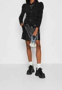 Nainen, jolla on yllä musta puffi-hihainen takki ja napitettu nahkahame, pitää kädessään pientä denimitaskua, valkoisia sukkia ja jykeviä mustia kenkiä.