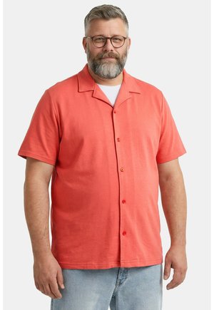 Mann mit grauem Bart und Brille, der ein korallenfarbenes kurzärmeliges Hemd über einem weißen T-Shirt und hellblauen Jeans trägt.
