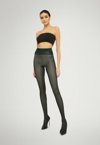 Collant verdi opachi con una ampia fascia nera in vita, abbinati a un top nero senza spalline e tacchi alti, caratterizzati da una texture liscia.