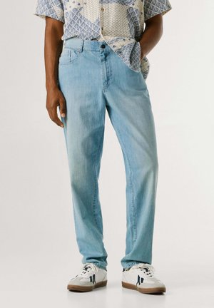 Homme portant un jean bleu clair, des baskets blanches avec des détails noirs, et une chemise à manches courtes à motifs, une main dans la poche.