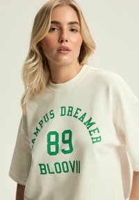 Kremowy, luźny sweatshirt z zielonym nadrukiem "CAMPUS DREAMER 89 BLOOVII." Miękka faktura, okrągły dekolt i krótkie rękawy.