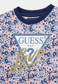 T-shirt con motivo floreale nelle tonalità di blu e rosa, caratterizzata da un logo triangolare blu con la scritta "GUESS" e un accento dorato glitterato con la parola "Love".