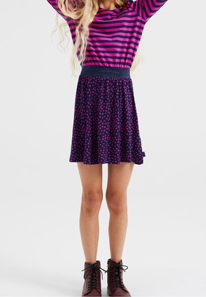Junge Person trägt einen marineblauen Rock mit rosa Punkten, ein rosa und marineblau gestreiftes langärmeliges Oberteil und dunkelrote Schnürstiefel.