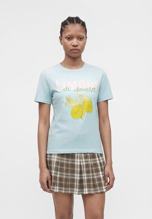 Młoda osoba z zaplecionymi włosami, ubrana w jasnoniebieski t-shirt z grafiką cytryny i napisem „Limoni di Amalfi”, w połączeniu z brązową, kratowaną spódnicą.