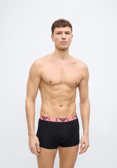 Άνδρας χωρίς πουκάμισο, όρθιος, φορώντας μαύρα μπόξερ με ροζ μέση που διαθέτει μαύρο λογότυπο και γράμματα, απλό ανοιχτόχρωμο φόντο.