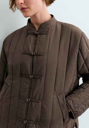 Femme portant une veste matelassée marron avec des coutures verticales et des fermetures par boutons en forme de grenouille traditionnels chinois, main dans la poche latérale.
