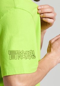 Neonově zelené tričko s krátkými rukávy a grafickým designem černými znaky na levém rukávu. Vyrobeno z bavlněné tkaniny.