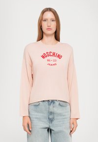 Pull long à manches longues rose clair, col rond, arborant le logo rouge "MOSCHINO 86 23 JEANS", texture côtelée, associé à un jean en denim bleu.