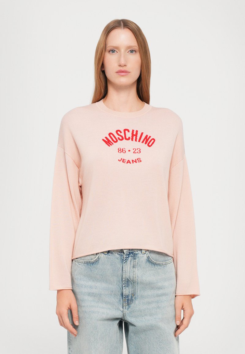 Pull long à manches longues rose clair, col rond, arborant le logo rouge "MOSCHINO 86 23 JEANS", texture côtelée, associé à un jean en denim bleu.