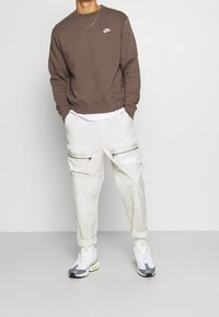 Muž v hnědé mikině Nike, bílé vrstvené košili, béžových cargo kalhotách Nike a bílých šedých teniskách Nike stojí s rukama v kapsách.