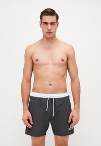 Bañadores de hombre en gris oscuro, con una cinturilla elástica blanca y cordón, y un logo blanco en el lado izquierdo. Textura suave.