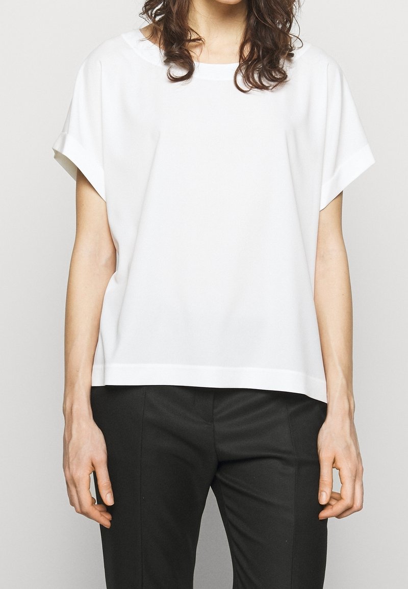 Personne portant un haut blanc uni à manches courtes et un pantalon noir, visible du cou jusqu'à mi-cuisse, debout sur un fond clair.