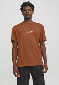 Camiseta de algodón marrón con un corte relajado, mangas cortas y cuello redondo. Logotipo "Originals Studio" impreso en blanco en el pecho.