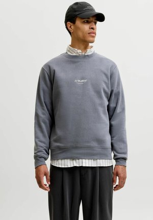 Homme portant un sweat-shirt gris JJ Studios par-dessus une chemise blanche à rayures, un pantalon noir et une casquette noire, regardant vers la droite sur un fond uni.