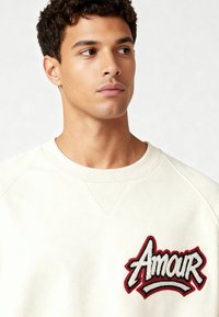 Sweat-shirt crème à col ras du cou et manches raglan. Doté d'un patch brodé "Amour" en rouge et noir à l'avant. Texture douce.