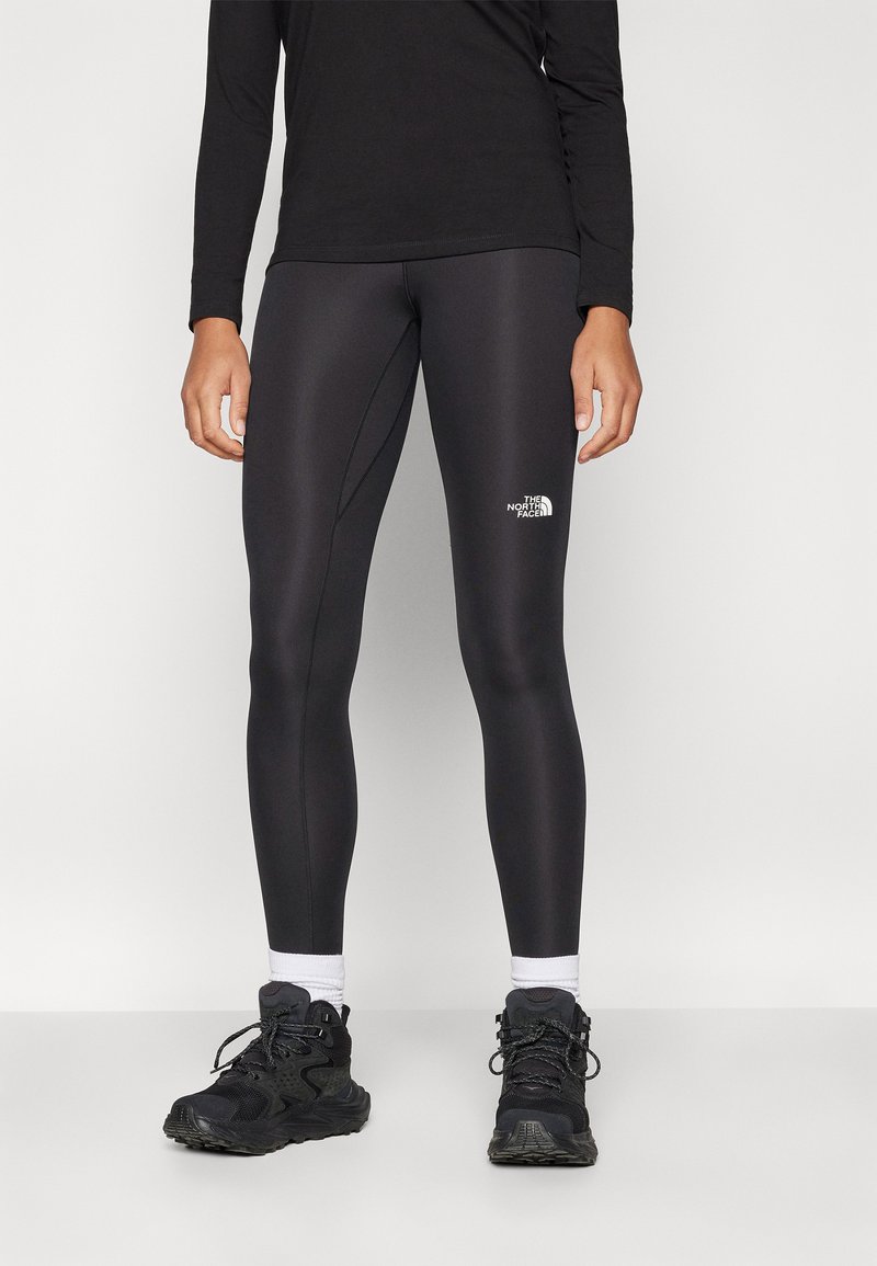 Leggings sportivi neri con una texture liscia e un design aderente, caratterizzati da un piccolo logo bianco sulla coscia superiore. Abbinati a sneakers nere.
