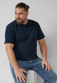 Homme aux cheveux attachés et à la barbe, portant un t-shirt bleu marine et un jean bleu, assis sur un bloc blanc, regardant vers la gauche.