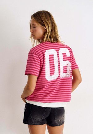 Frau mit schulterlangem blondem Haar, die ein pink gestreiftes T-Shirt mit weißer Nummer 06 und schwarze Denim-Shorts trägt, seitlich betrachtet.