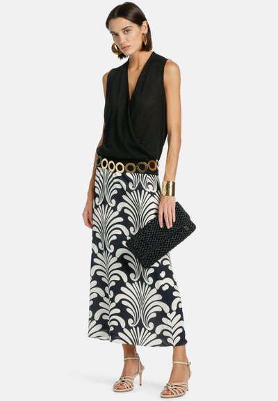 Femme portant un haut noir sans manches, une jupe à motif noir et blanc, une ceinture dorée et un bracelet manchette, tenant une pochette tissée noire, sandales à talons beiges.