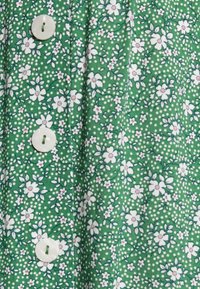 Groene stof met een bloemenpatroon van witte en roze bloemen, omringd door kleine witte stippen. Drie grote witte knopen zichtbaar.
