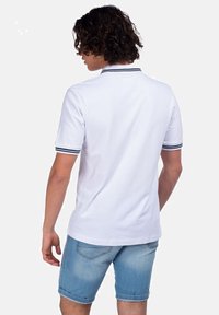 Polo bianco con colletto e polsini a righe navy, realizzato in tessuto liscio. Indossato con pantaloncini in denim chiaro, presenta orli arrotolati.
