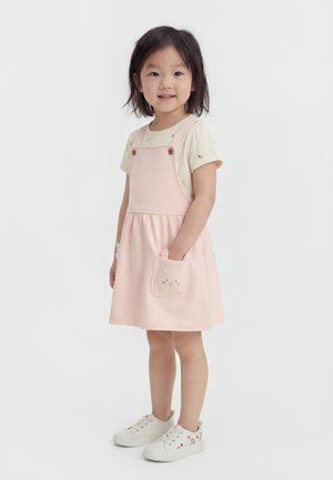 Young girl wearing a light pink pinafore dress with cat face pockets, a cream t-shirt, and white sneakers with floral embroidery.