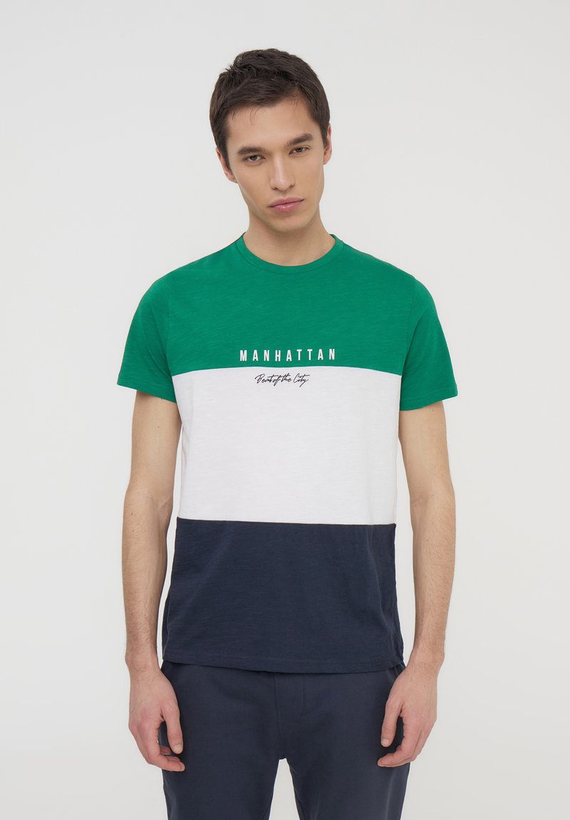T-shirt a blocchi di colore verde, bianco e blu navy. Presenta un colletto girocollo e la scritta stampata "MANHATTAN" sul petto. Maniche corte.