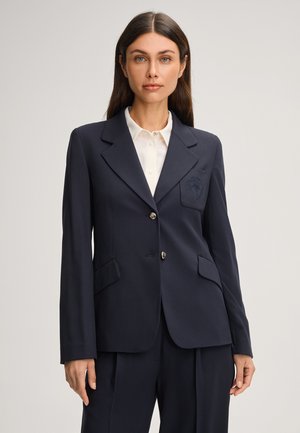 Navy tailored blazer with two buttons, notch lapels, and side pockets. Features an embroidered patch on the left chest pocket. Smooth fabric.