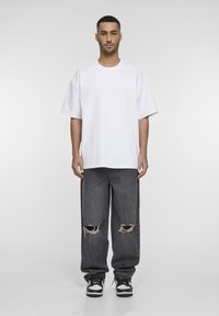 Hvid oversized T-shirt, mørkegrå baggy jeans med slidte huller, og sorte/hvide sneakers. Simpelt design, naturlig stoftekstur.