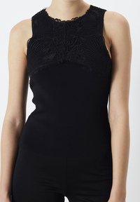 Bezrękawnik w kolorze czarnym z teksturowanym koronkowym wykończeniem na górnej części klatki piersiowej i ramionach, noszony z czarnymi dopasowanymi spodniami.