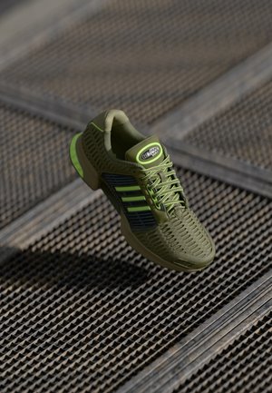 Chaussure de sport verte avec des bandes noires et vert fluo et le logo "Climacool", positionnée au-dessus d'une grille métallique texturée.