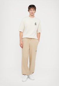 Beige cotton T-shirt with short sleeves and a black logo, paired with beige relaxed sweatpants featuring a side pocket and black lettering. White sneakers.