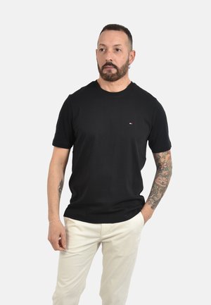 Uomo con la barba e il braccio destro tatuato che indossa una camicia nera a maniche corte e pantaloni bianchi, in piedi con la mano sinistra in tasca.