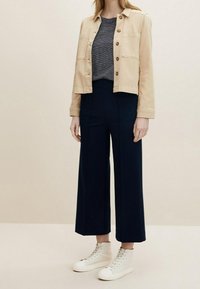 Kvinna i en beige knäppt jacka, svart och vit randig topp, marinblå vida byxor och vita högklackade sneakers mot en enfärgad bakgrund.