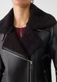 Zwarte leren jas met een textuurrijke zwarte shearlingkraag, asymmetrische rits en zijzakken. Close-up benadrukt de details.