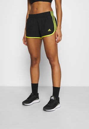 Kobieta w czarnych sportowych szortach Adidas z fluorescencyjnym żółtym obszyciem, czarnych skarpetach i czarnych butach do biegania Adidas, stojąca na szarej podłodze.