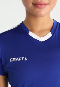Blå sportsjersey med hvide detaljer. Har et V-neck design og "CRAFT" logoet på venstre side. Fremstillet af åndbart stof.