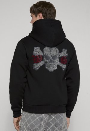 Mand iført sort hættetrøje med nitte-hoved og korsben design og røde bogstaver "D" og "S" på ryggen.